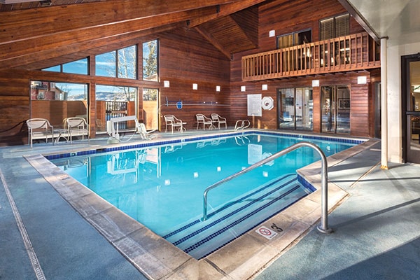An indoor swimming pool is framed by large windows, allowing natural light to illuminate the space. The pool is surrounded by lounge chairs, and a wooden ceiling enhances the warm atmosphere. A safety railing provides access to the water.
