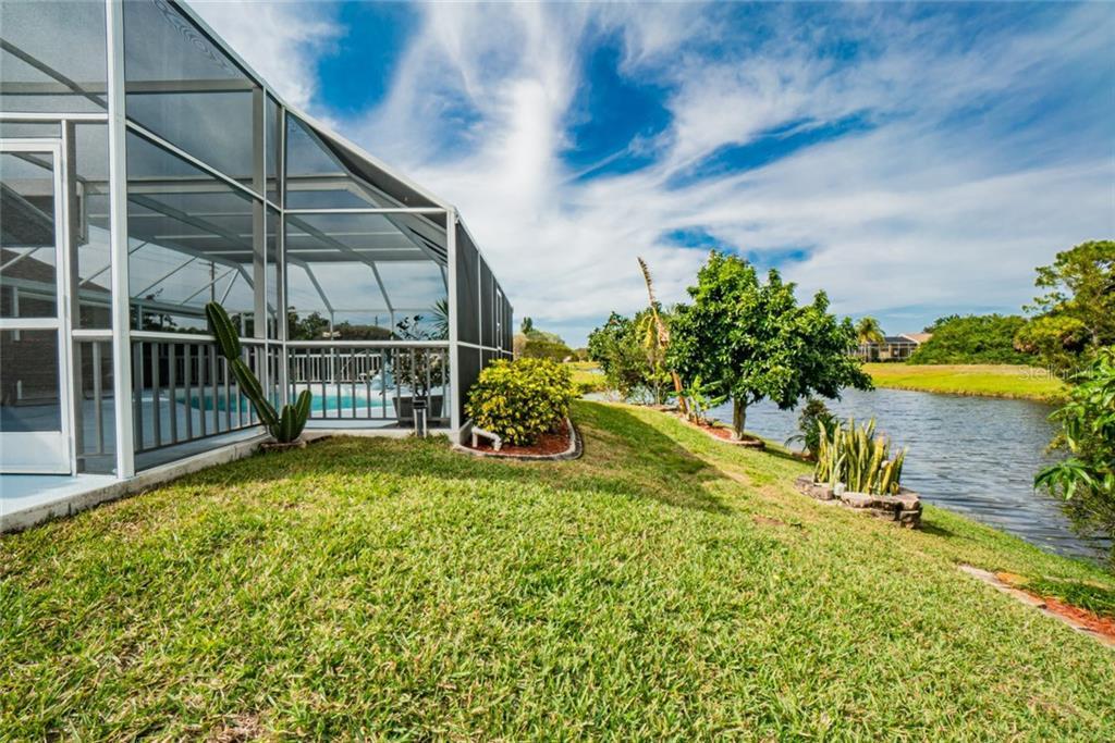 A grassy area lines the edge of a canal, with the water visible alongside the property. Lush greenery, including shrubs and small trees, adds natural beauty. The screened lanai structure is seen in the background, featuring expansive views of the serene surroundings.