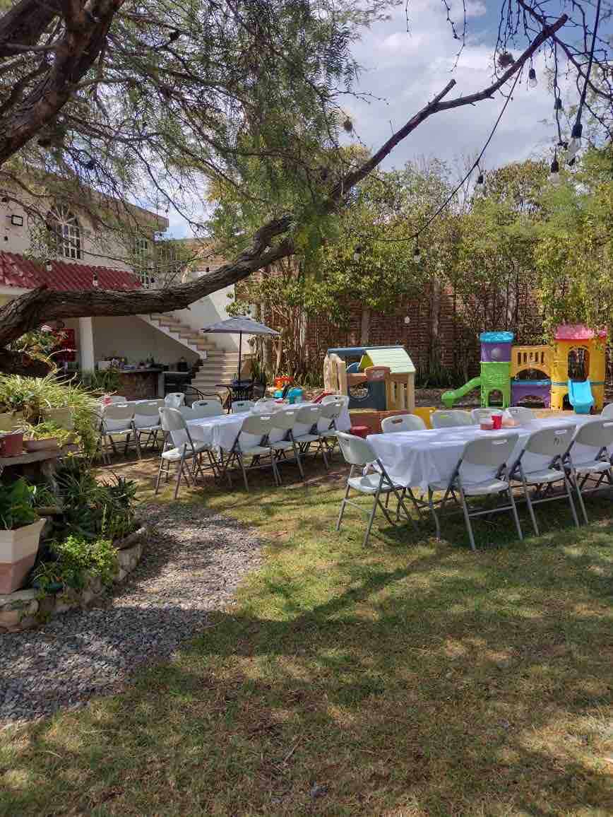 A spacious outdoor area is presented, featuring a collection of folding tables surrounded by chairs. A children's play structure is visible, alongside a well-maintained garden area. Lush greenery and a climbing tree create a natural setting, with a glimpse of a building in the background.