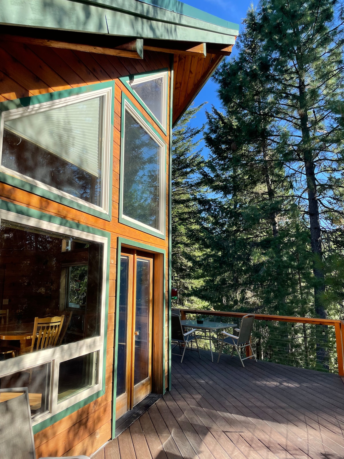 The exterior of the cabin is shown, featuring large windows that invite natural light, complemented by wooden siding and a green roof. A spacious deck is visible, with patio furniture arranged for outdoor relaxation amid tall trees.