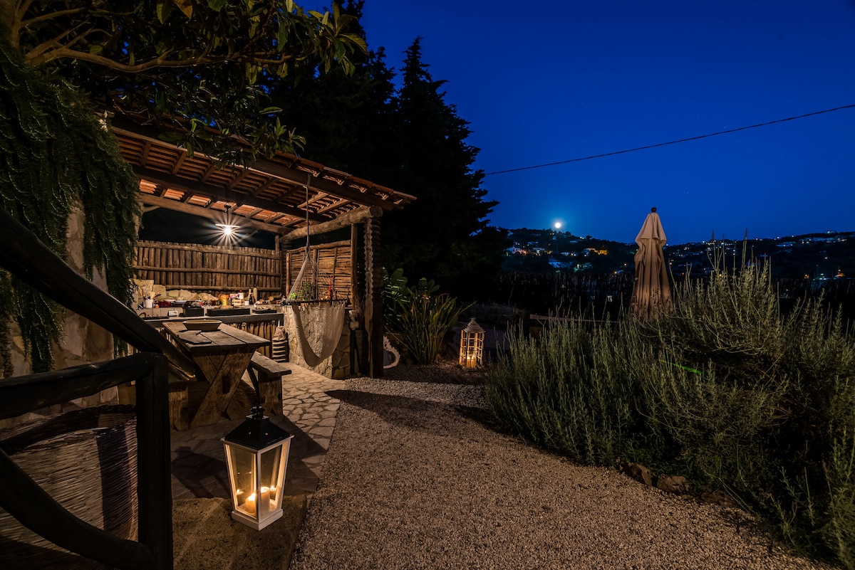 An outdoor kitchen area is featured under a wooden pergola, illuminated by hanging lights. A rustic table is set among the gravel pathway, surrounded by plants and lanterns. Nighttime scenery reveals distant lights twinkling over the hillside, enhancing the serene atmosphere.