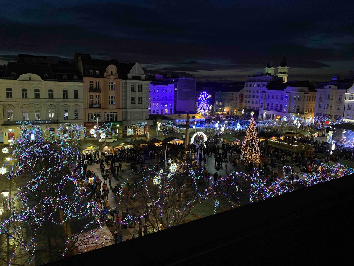 A vibrant view of a holiday market is displayed, illuminated by colorful lights amidst tall trees. A prominent Christmas tree stands in the center, surrounded by stalls and visitors enjoying the festive atmosphere, with a Ferris wheel and historical buildings enhancing the lively scene.