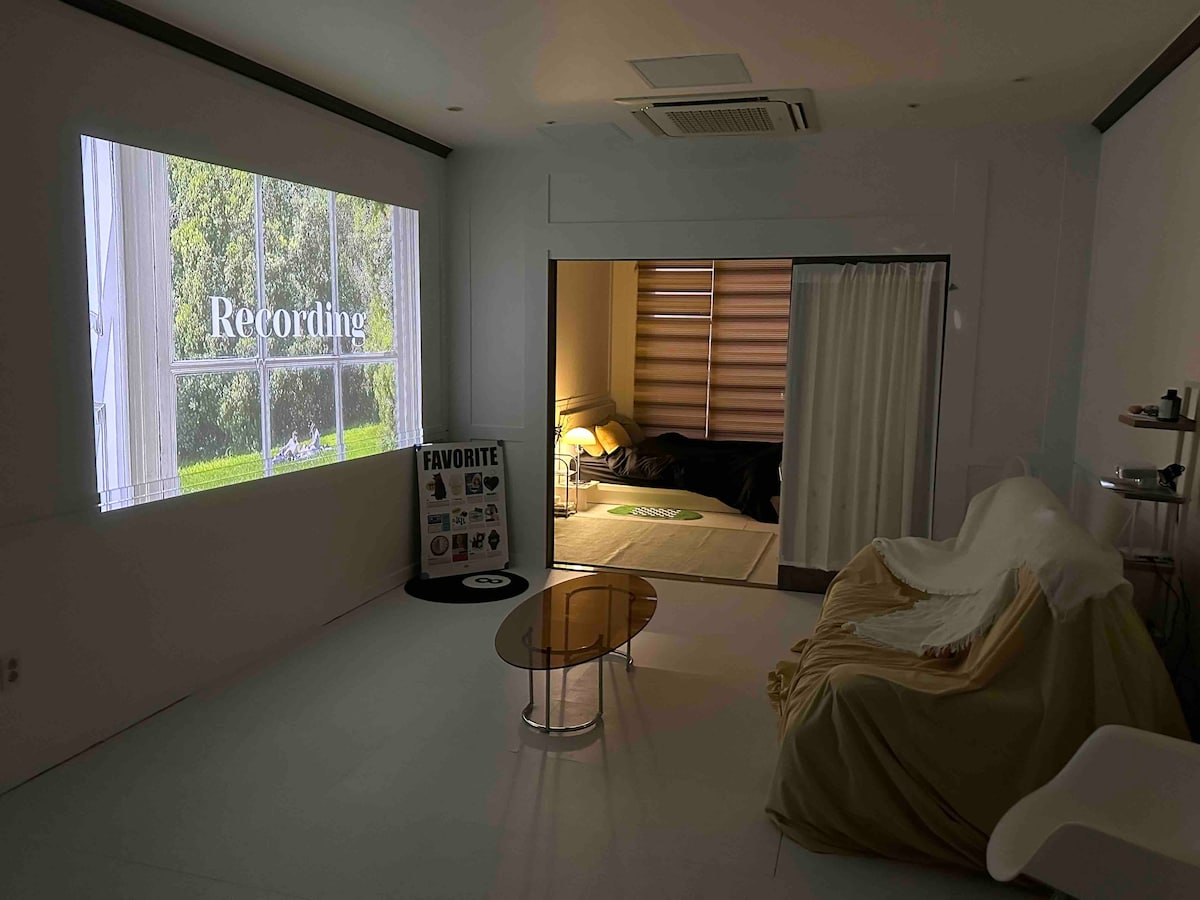 A spacious room is depicted, featuring a large projection screen displaying 'Recording' alongside a cozy seating area with a light-colored blanket draped over a sofa. A partition provides a glimpse of a sleeping area with a bed, subtly inviting relaxation.