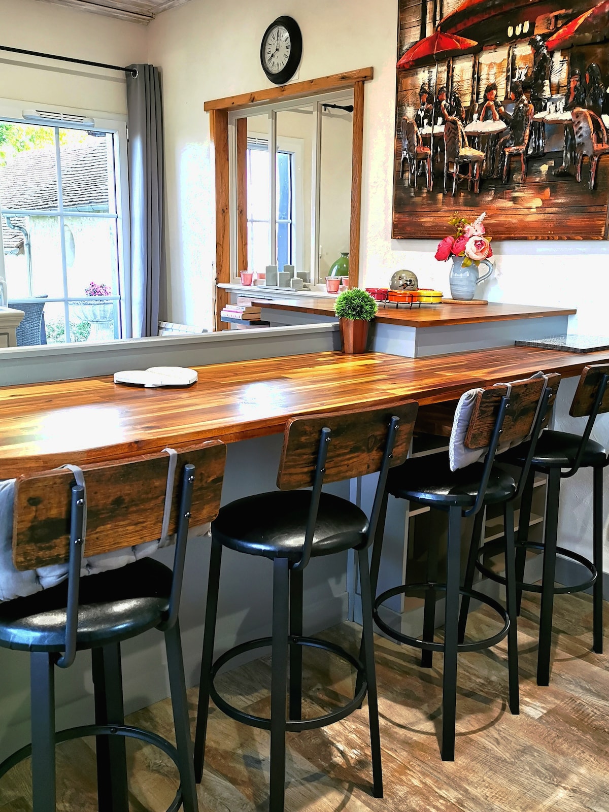 The kitchen bar features a long wooden countertop supported by modern stools. A large mirror reflects the natural light from nearby windows, enhancing the space. An artwork depicting a dining scene adds character to the wall, creating a functional and welcoming atmosphere.