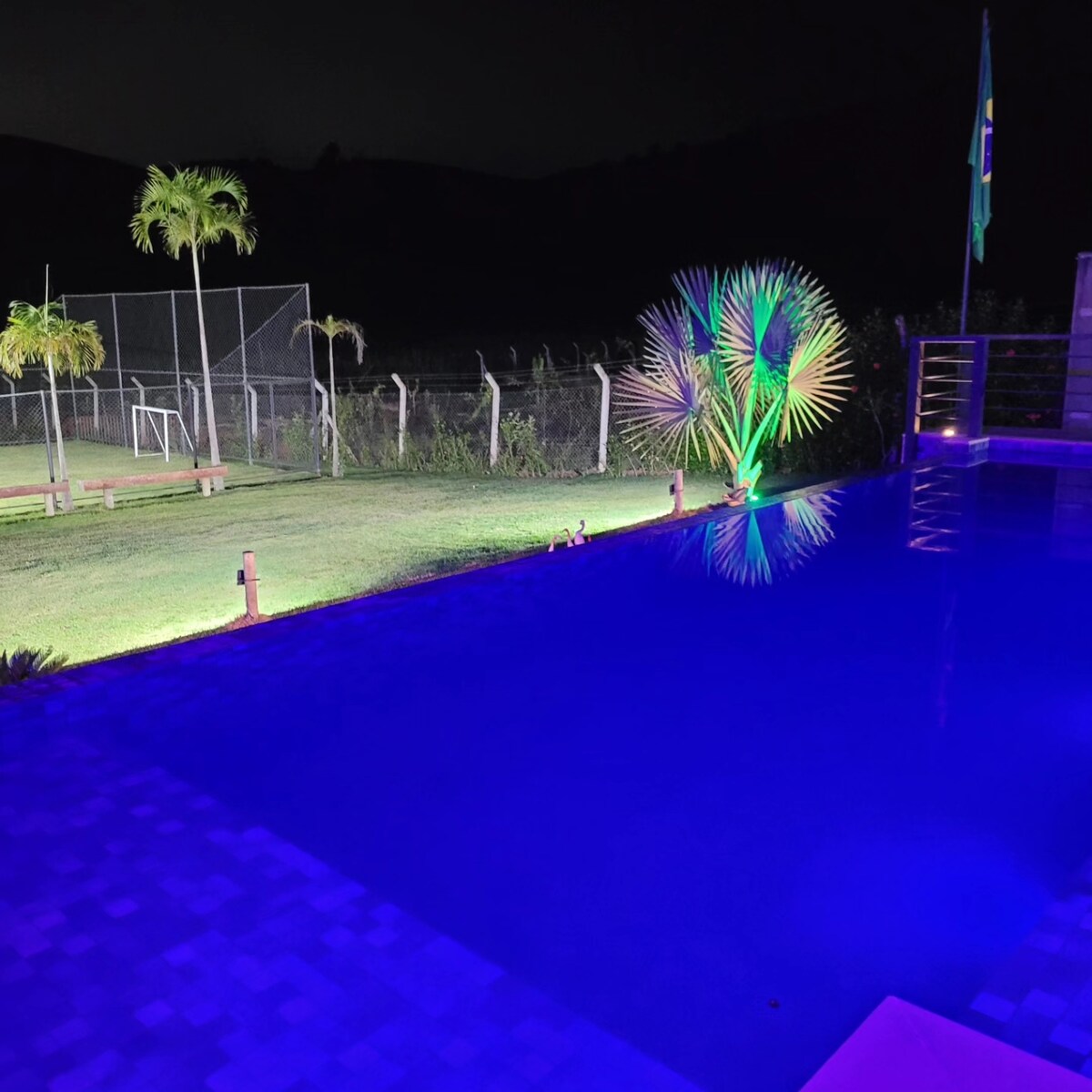The image showcases an illuminated infinity pool reflecting vibrant blue lights at night. Lush greenery and decorative palm plants are positioned around the pool area. A sports field with goalposts is visible in the background, enhancing the outdoor recreational space.