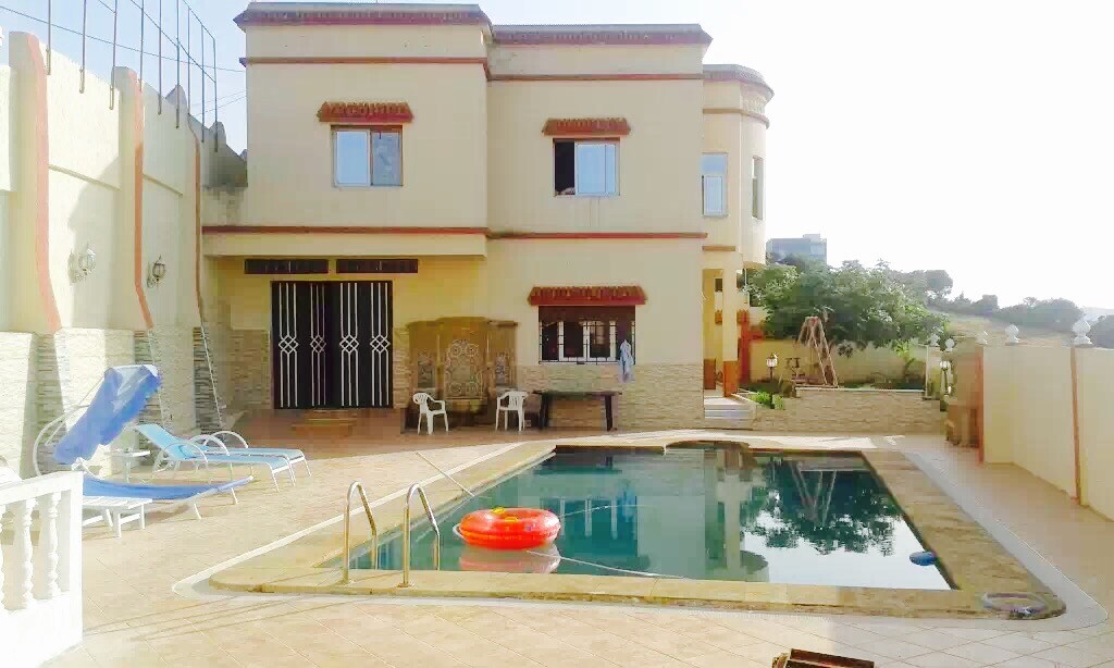 A tranquil outdoor area features a swimming pool surrounded by tiled flooring. Several lounge chairs are positioned nearby, and a colorful inflatable ring floats in the water. The villa's exterior showcases multiple windows and a shaded patio space, with well-maintained greenery in the background.