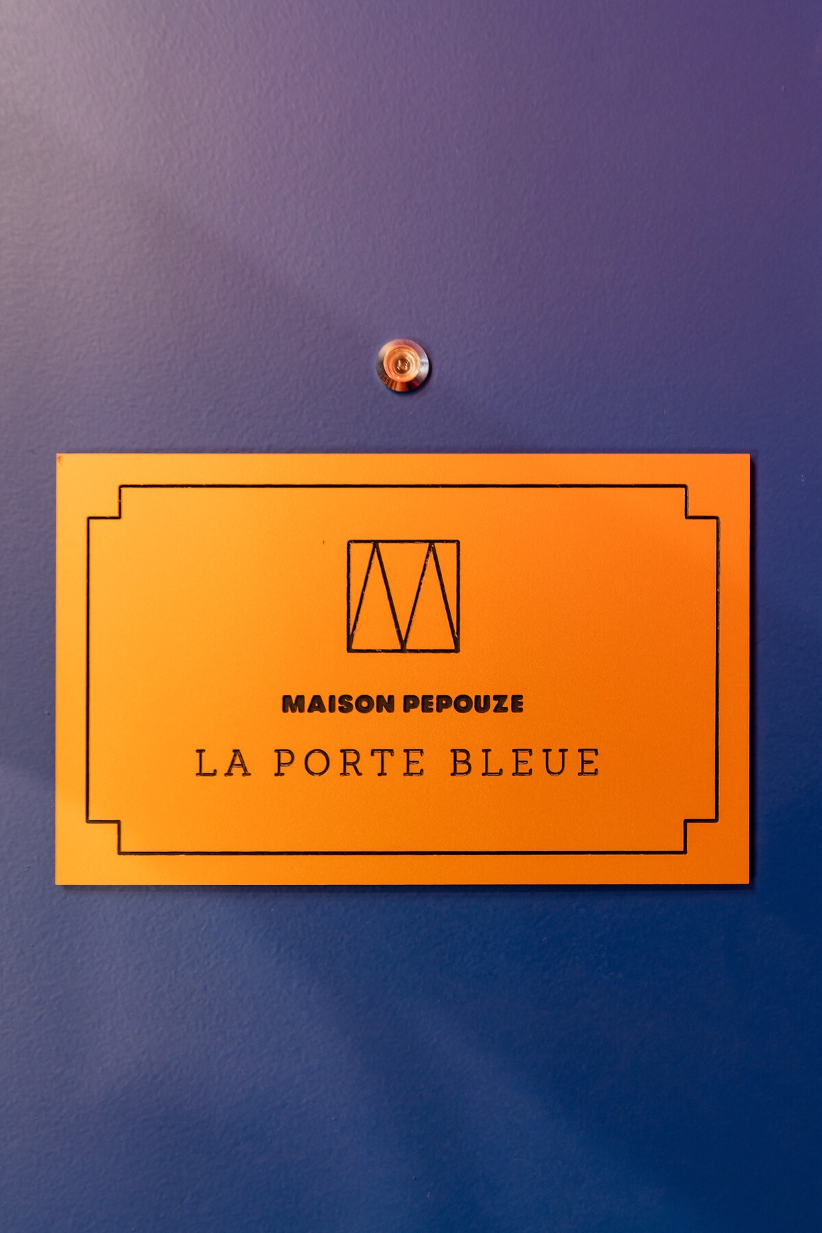 A vibrant orange sign is mounted on a deep blue door, displaying the name 'La Porte Bleue' in bold lettering. The sign is accented with an elegant logo at the top, adding a touch of sophistication to the entrance.