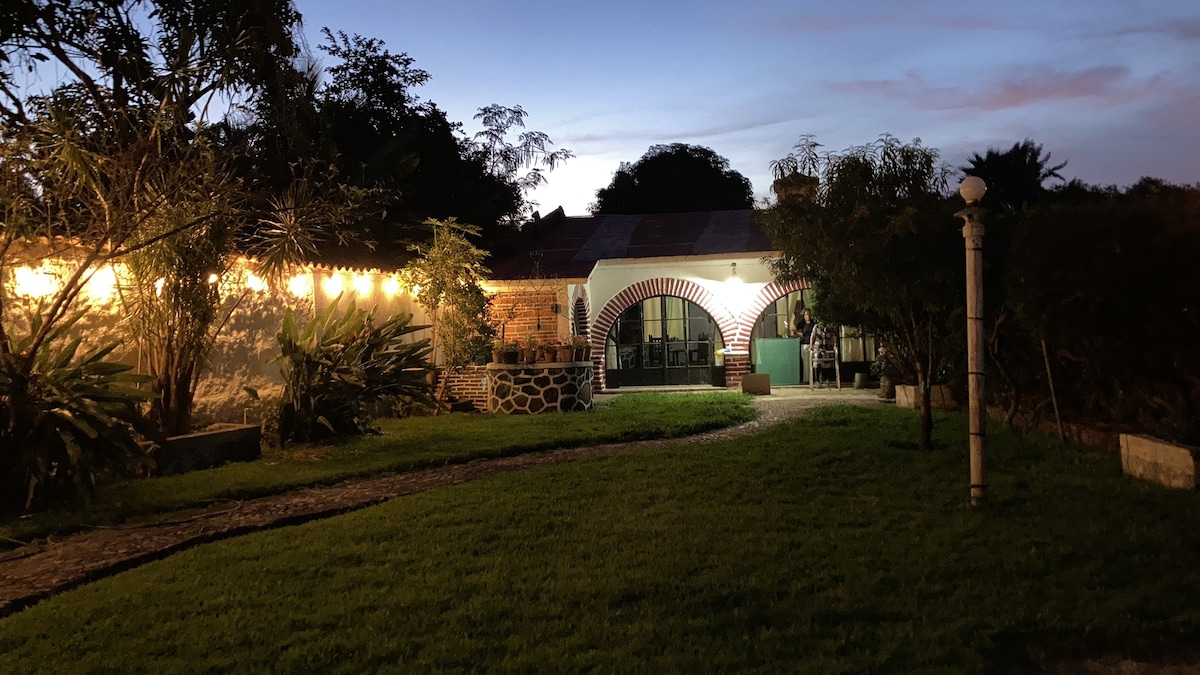 A welcoming garden space is illuminated by soft lights at dusk. The entryway features a stone pathway leading to an arched façade, framed by lush greenery and shrubs. The structure showcases expansive windows, inviting glimpses of the interior that complements the tranquil outdoor ambiance.