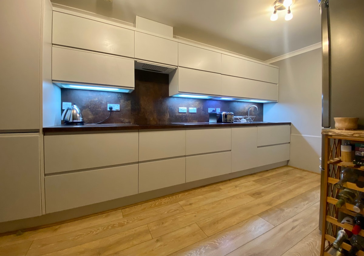 The kitchen features modern cabinetry with sleek lines and a contrasting dark countertop. Under-cabinet lighting enhances the workspace. Essential appliances, including a kettle and coffee machine, are visible, while a wood floor adds warmth to the area.