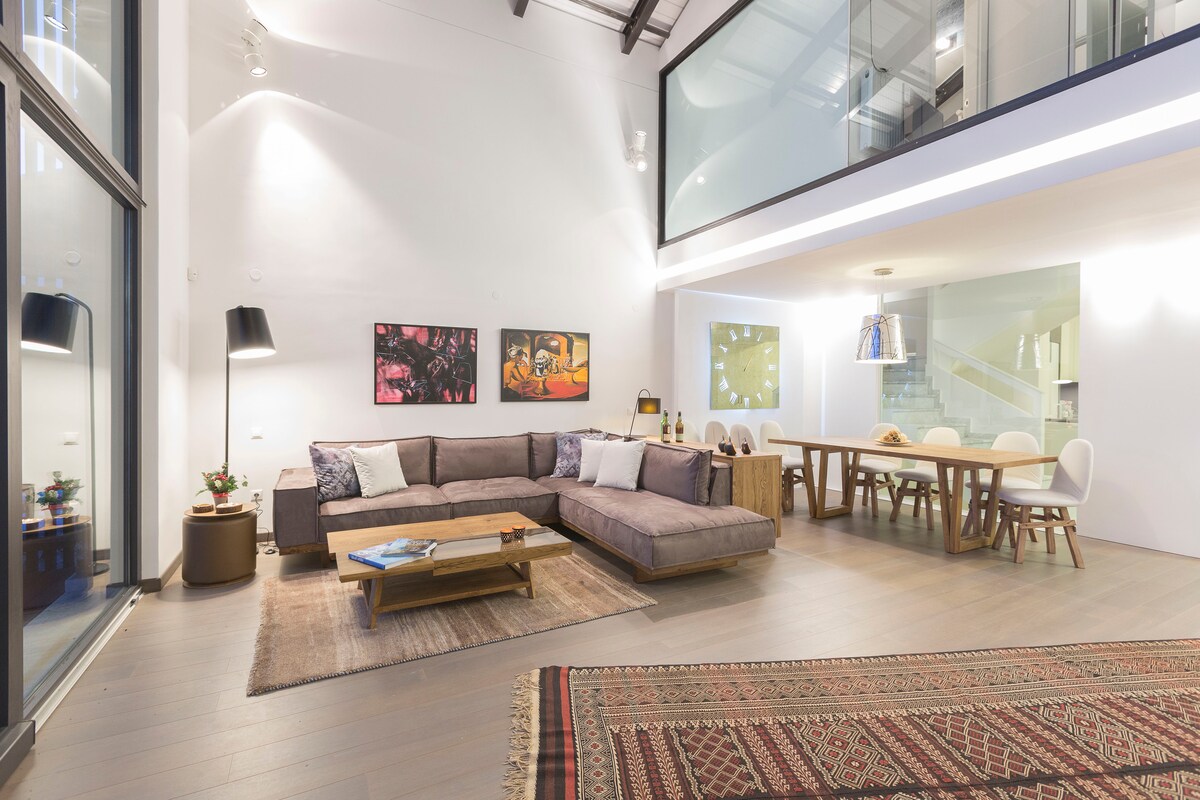 A spacious living area features a large sectional sofa adorned with decorative pillows, complemented by a wooden coffee table. Colorful wall art adds vibrancy, while a dining table with white chairs is illuminated by a pendant light. Natural light enters through expansive windows, enhancing the bright ambiance.