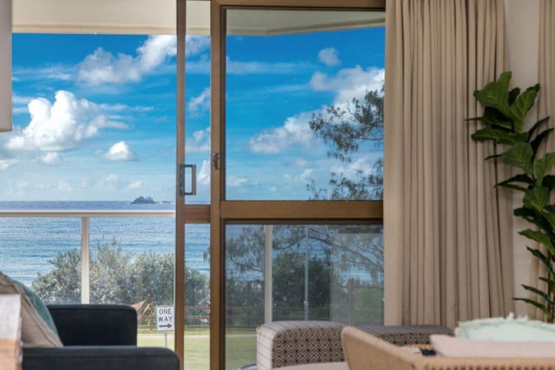A glass sliding door frames a serene ocean view, with gentle waves and a distant island visible. Soft curtains are drawn to the sides, and hints of green foliage can be seen outside, creating a sense of openness in the living area.