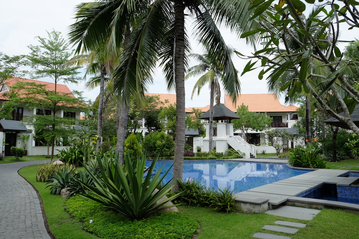 The landscaped grounds feature a serene swimming pool surrounded by lush tropical vegetation. Palm trees and well-kept gardens create a relaxing environment. Pathways lined with greenery lead toward various villa structures in the background, enhancing the tranquil atmosphere of the resort.