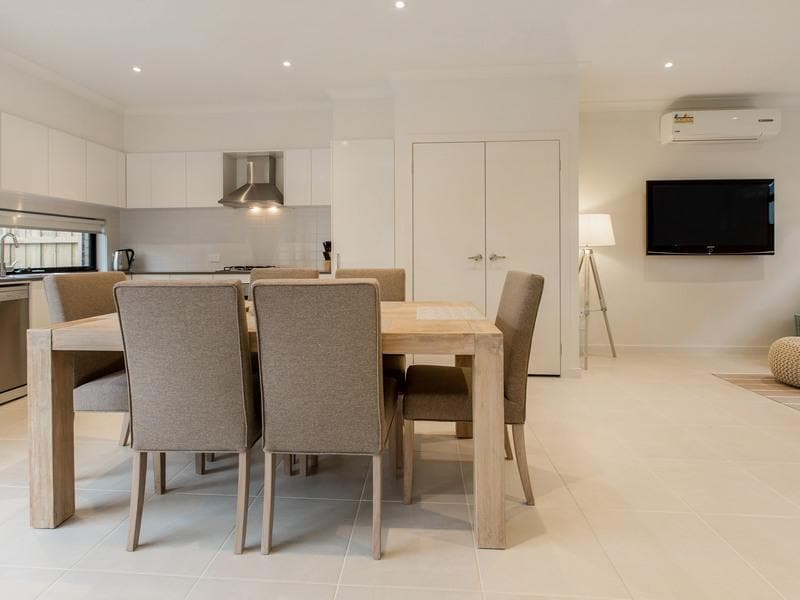 The open-plan kitchen and dining area features a modern wooden dining table surrounded by six upholstered chairs. A well-equipped kitchen is visible in the background, alongside a wall-mounted TV. Soft lighting and pale tile flooring provide a bright and clean ambiance.