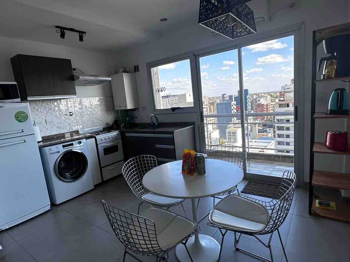 A modern kitchen and dining area is presented, featuring a round table with white wire chairs. Large windows provide a view of the city, while an equipped kitchen includes a stove, refrigerator, and washing machine. Natural light fills the space, enhancing the clean design.