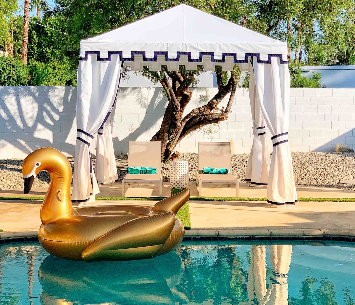 A spacious pool area features a golden inflatable swan floating on clear water. A shaded cabana with elegant drapery provides seating, accompanied by two lounge chairs. Lush greenery and a nearby tree enhance the outdoor ambiance, creating a relaxing retreat.