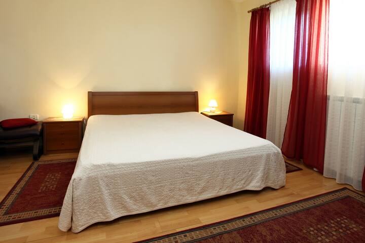Quarto iluminado com uma grande cama de casal, roupa de cama branca, abajures aconchegantes, móveis de madeira, cortinas vermelhas, piso de parquet e estilo clássico simples.
