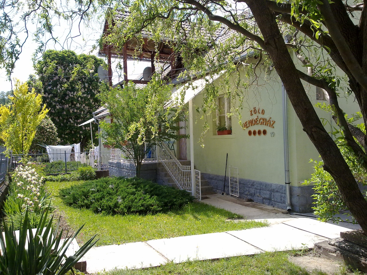 A charming guesthouse is surrounded by a lush garden with green shrubs and trees. A paved walkway leads to the entrance, where a sign indicates the name of the accommodation. Sunlight filters through the leaves, creating a serene atmosphere.