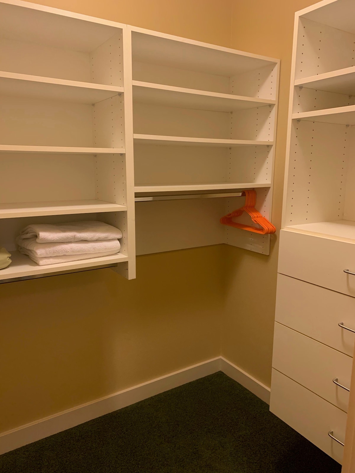 A walk-in closet is displayed, featuring built-in shelving and a drawer unit. Fresh white towels are neatly folded on a shelf, while a bright orange hanger is visible on a clothing rod. The walls are painted in a soft, warm hue.
