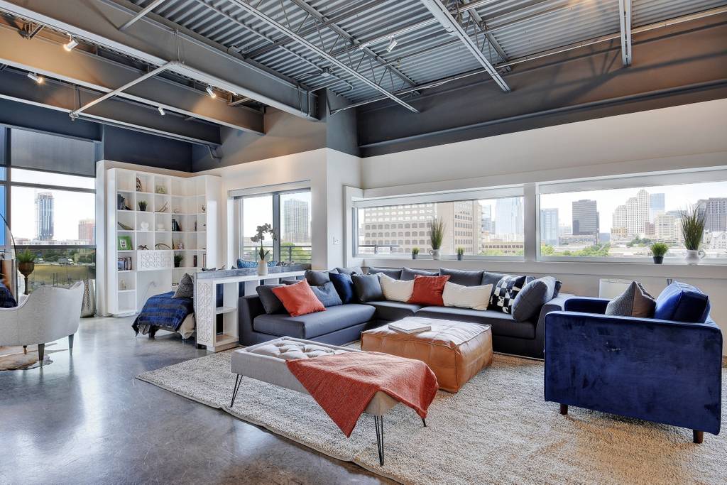 The spacious living area features large windows allowing natural light and city views. A sectional sofa and additional seating surround a coffee table, complemented by a textured area rug. Modern decor and shelving units enrich the ambiance, offering a blend of comfort and style.
