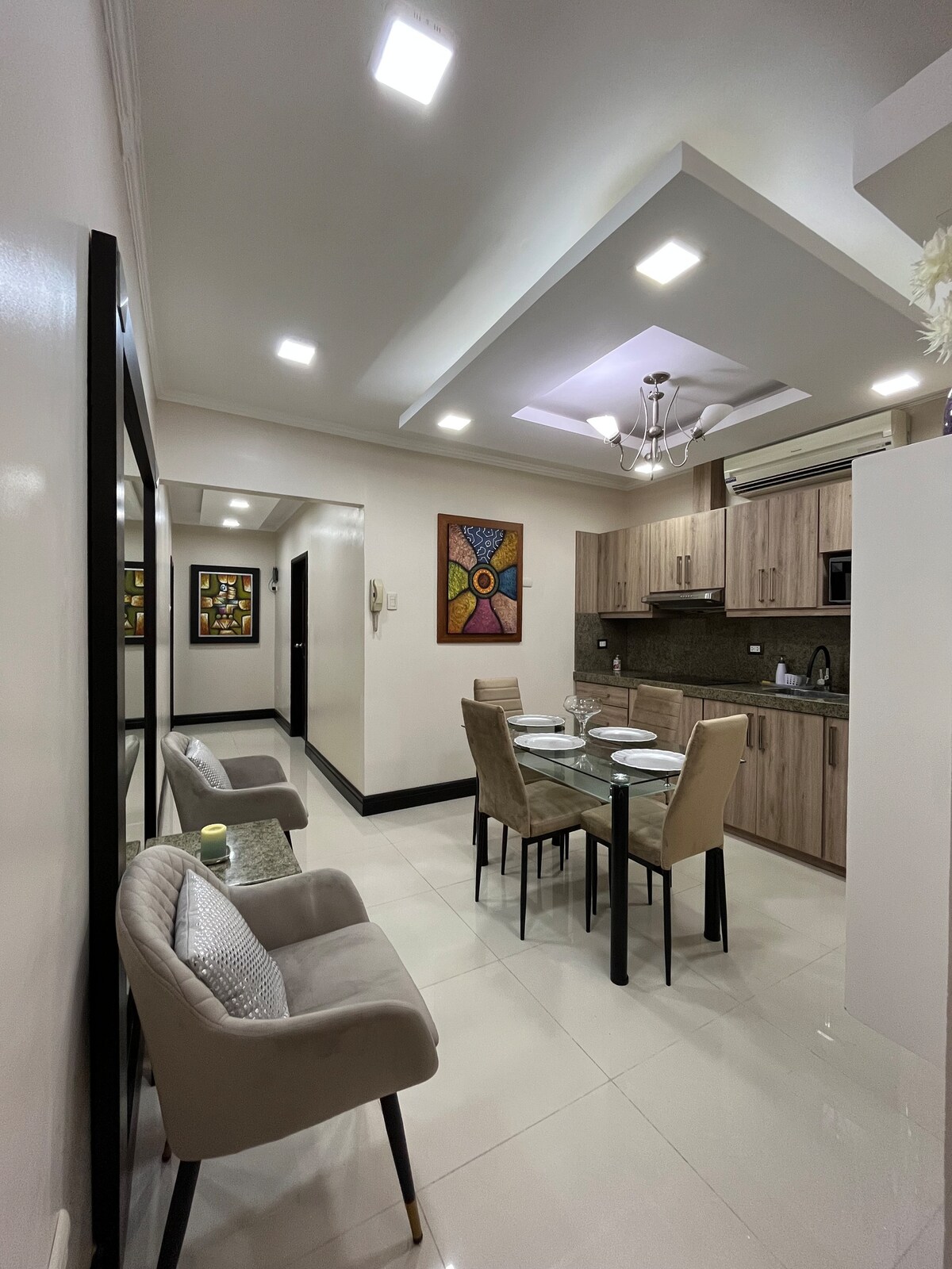 A modern kitchen and dining area are depicted, featuring a glass dining table surrounded by four chairs. The kitchen, with wooden cabinetry, includes appliances and counter space. Artwork is visible on the walls, complementing the bright lighting from the ceiling fixtures.
