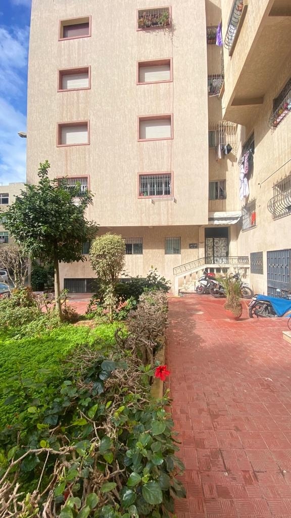 The building exterior presents a traditional Moroccan style, with a beige façade featuring several windows. A small garden area is visible, showcasing greenery and flowering plants. A pathway leads to the entrance, flanked by shrubs and bicycles parked nearby, contributing to a serene atmosphere.