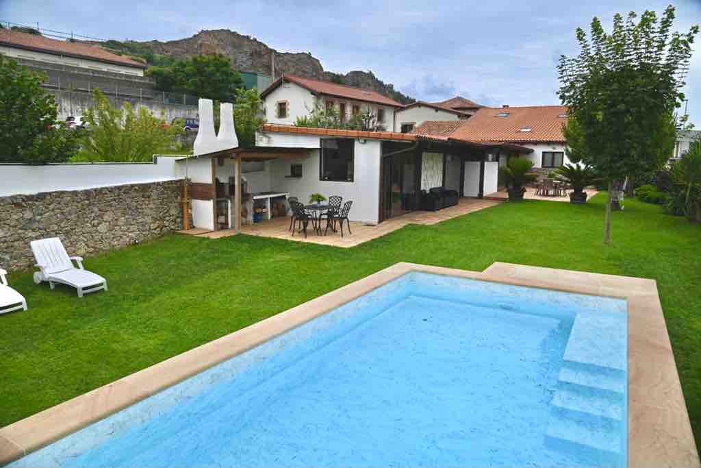 An outdoor area features a clear swimming pool with gentle steps leading in, surrounded by a lush green lawn. Lounge chairs are positioned nearby, and a patio with a dining table and chairs is visible, sheltered under a roof. The setting provides a view of the house and nearby hills.