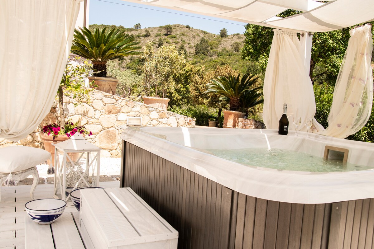 A private outdoor jacuzzi is surrounded by flowing drapes, offering a serene atmosphere. Potted plants and lush greenery create a tranquil backdrop, while a bottle of champagne rests nearby. The wooden decking adds a natural touch, inviting relaxation in a peaceful setting.