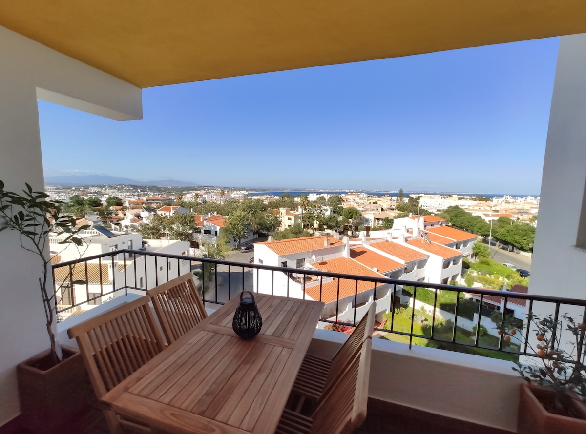 A balcony area features a wooden dining table with matching chairs, overlooking the expansive view of the surrounding neighborhood and distant coastline. Clear blue skies enhance the scenic backdrop, while a lantern adds a subtle touch of warmth. The railing provides safety while maintaining the view.