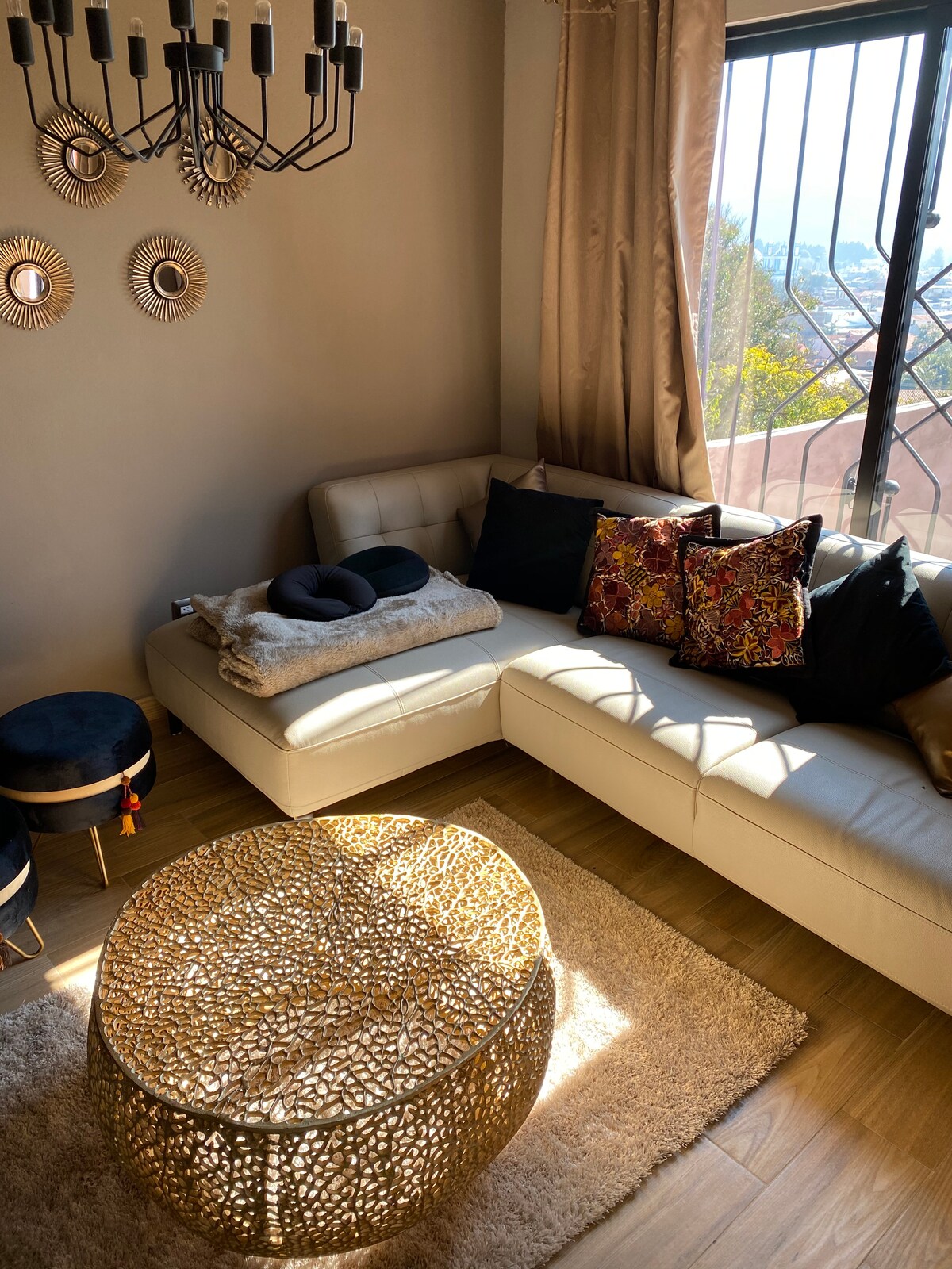 A light-filled living area features a modern beige sectional sofa adorned with decorative pillows in rich colors. A textured golden coffee table complements the space, while a plush rug adds comfort underfoot. Wall decor includes round mirrors that reflect natural light.