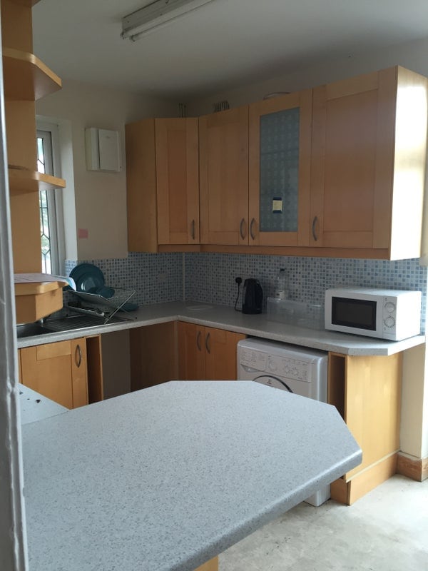The kitchen features light wood cabinetry with a tile backsplash. A countertop extends into a small breakfast bar area. Appliances include a microwave, kettle, and a washing machine, with additional dishware visible in the cabinets. Natural light filters in through a window.
