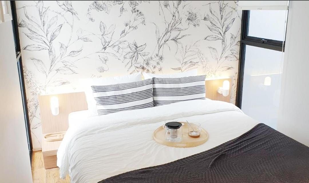 A well-lit bedroom features a spacious bed adorned with striped pillows and a soft blanket draped over the edge. A wooden tray with a drink rests on the bed, complemented by a floral wallpaper that adds a touch of nature to the atmosphere.
