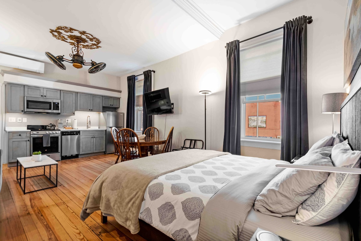 A spacious studio features a queen bed with patterned linens and plush pillows, complemented by large windows adorned with dark curtains. A dining area with a wooden table and chairs is visible, along with a fully equipped kitchen showcasing stainless steel appliances.