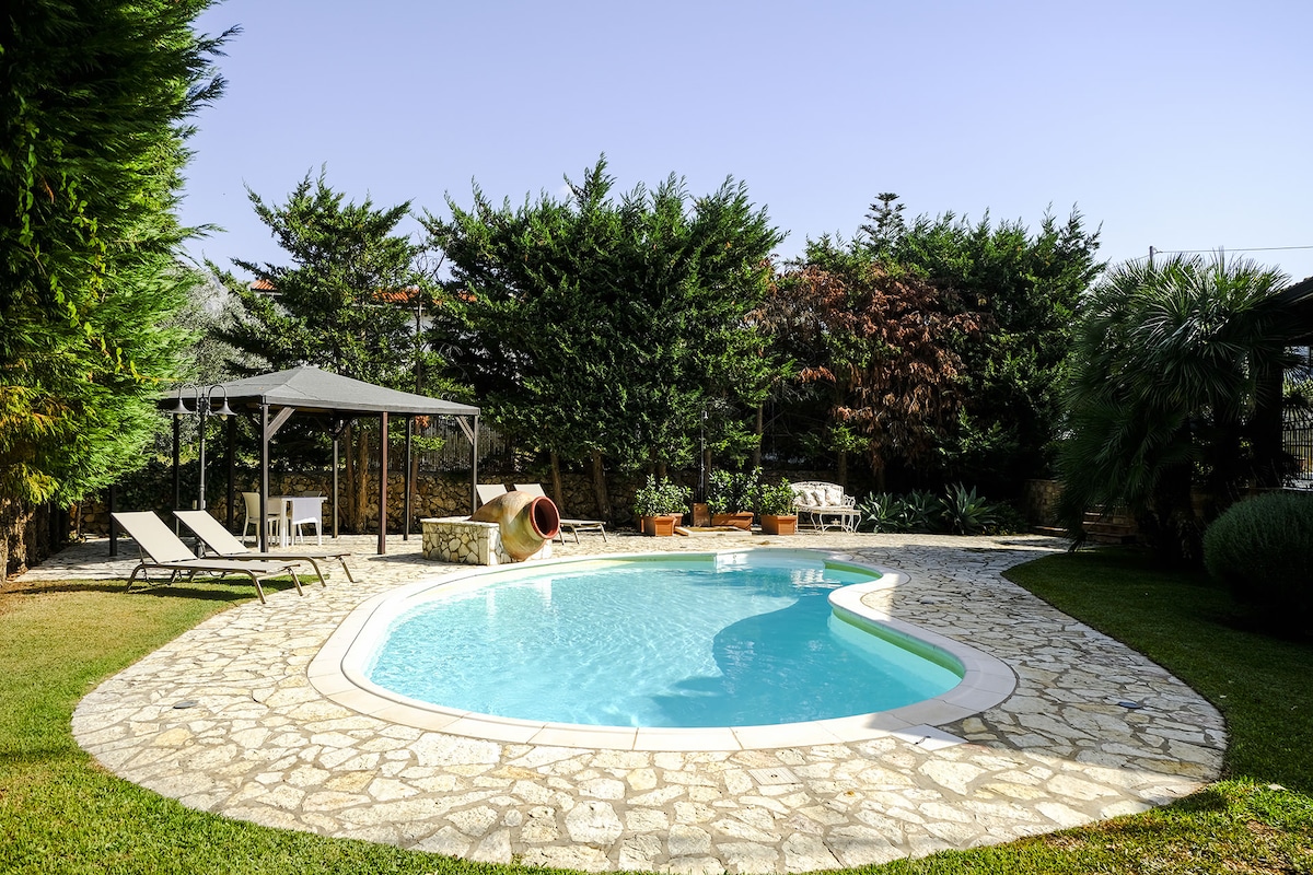 A curved pool is surrounded by a stone pathway and lush greenery, providing a serene outdoor space. Comfortable lounge chairs and a shaded area are nearby, offering opportunities for relaxation. Various plants and trees create a natural backdrop, enhancing the tranquil environment.