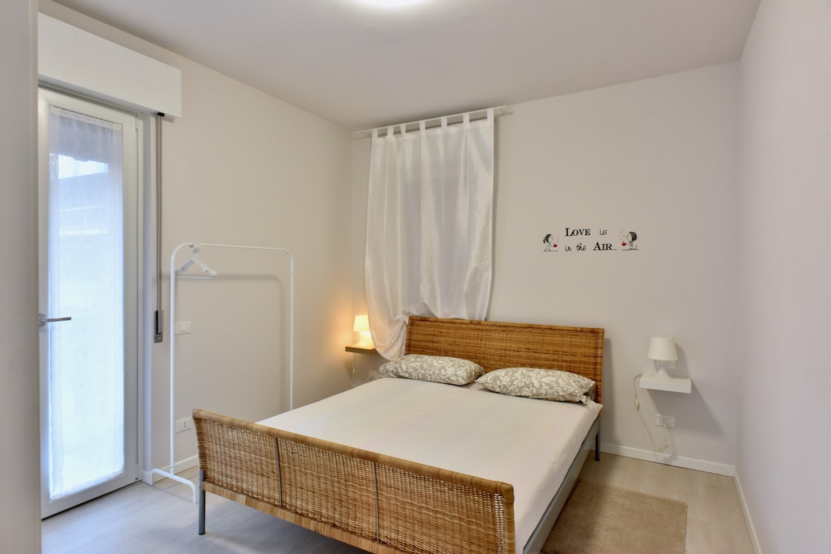 The calming bedroom features a spacious bed with soft, patterned pillows. A white curtain softly drapes over a glass door that opens to a small outdoor area. Minimalistic bedside lighting is provided by two wall-mounted lamps. A simple clothing rack adds functionality to the space.