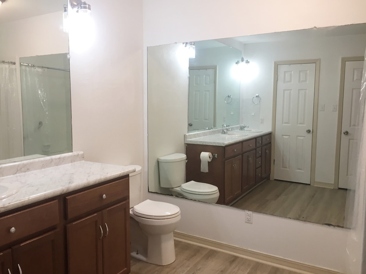 A spacious bathroom is featured with a large mirror reflecting the area. Light wood cabinetry provides storage space beneath a marble countertop. A toilet and a towel holder are positioned nearby. Light-colored flooring completes the clean and functional aesthetic.