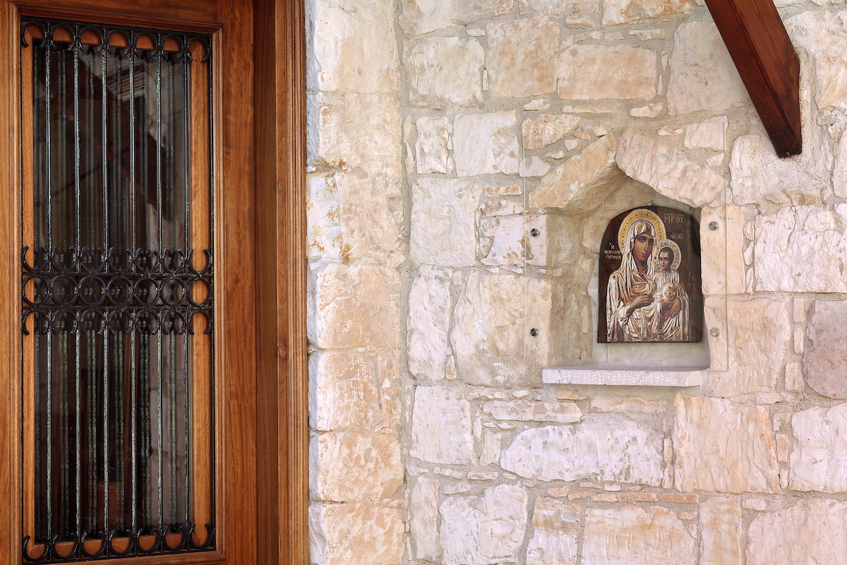 A rustic stone wall is featured, with a dark wooden door adorned with intricate iron detailing. To the right, an icon of a holy figure is placed within a stone niche, highlighting the home’s cultural heritage.