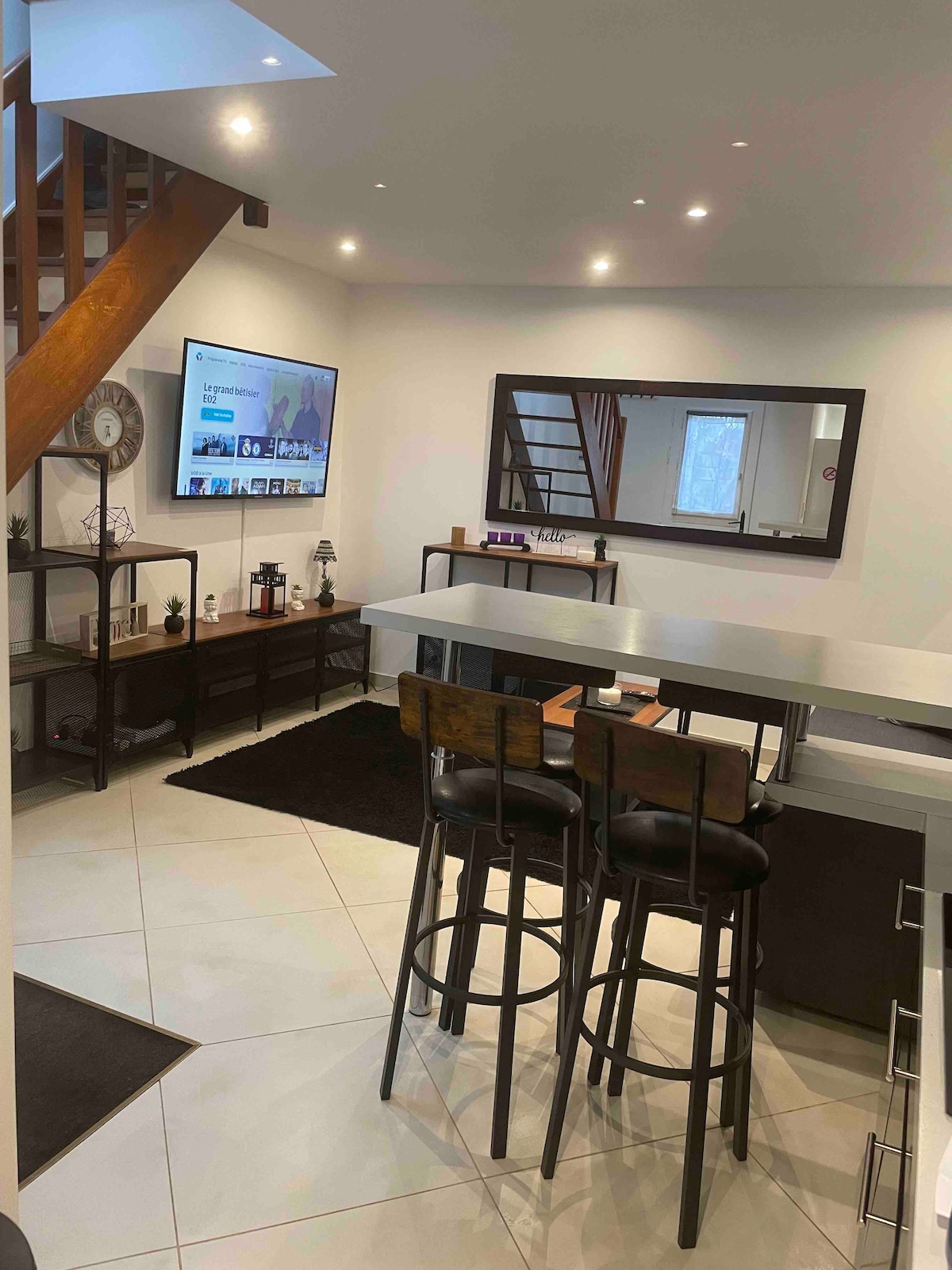 A modern living space features a sleek bar area with two high stools. A large wall-mounted television is visible above a minimalist media console. Bright lighting highlights the neutral tile flooring, while a nearby staircase adds vertical interest to the room.