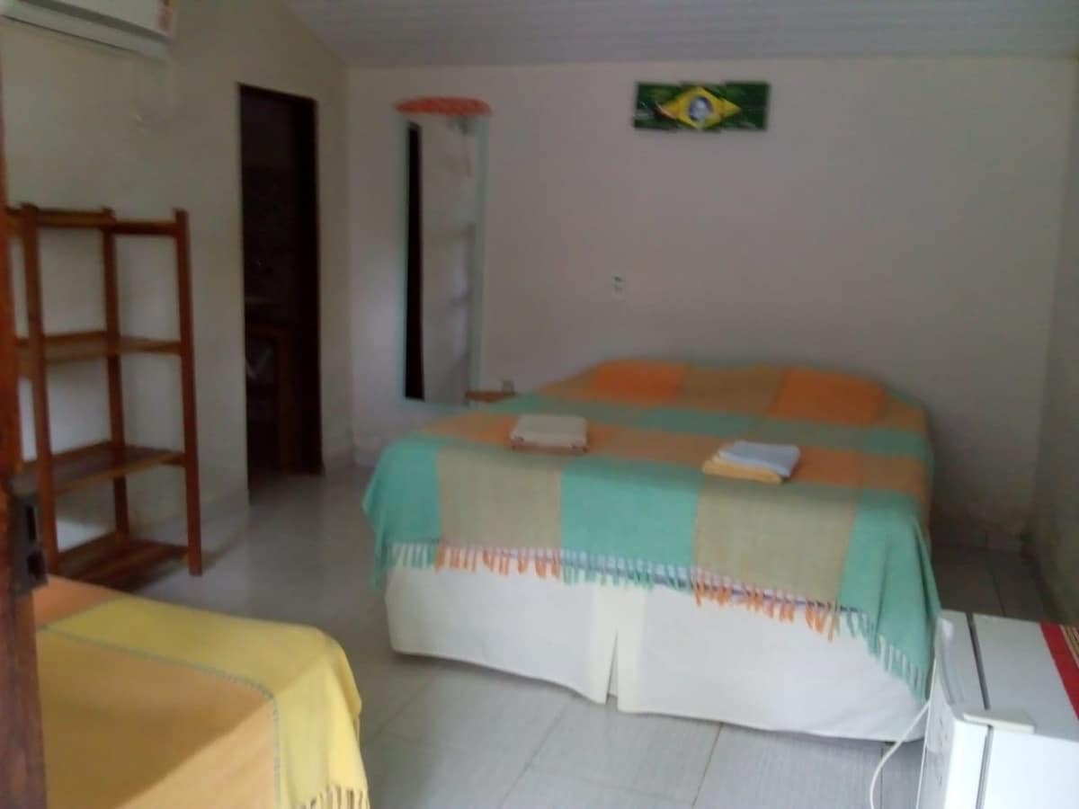 A bright bedroom features a spacious bed covered with a light blanket in pastel colors. A smaller bed is situated nearby, complemented by towels that are neatly arranged. A wooden shelf is placed against the wall, and a door leads to a private area.