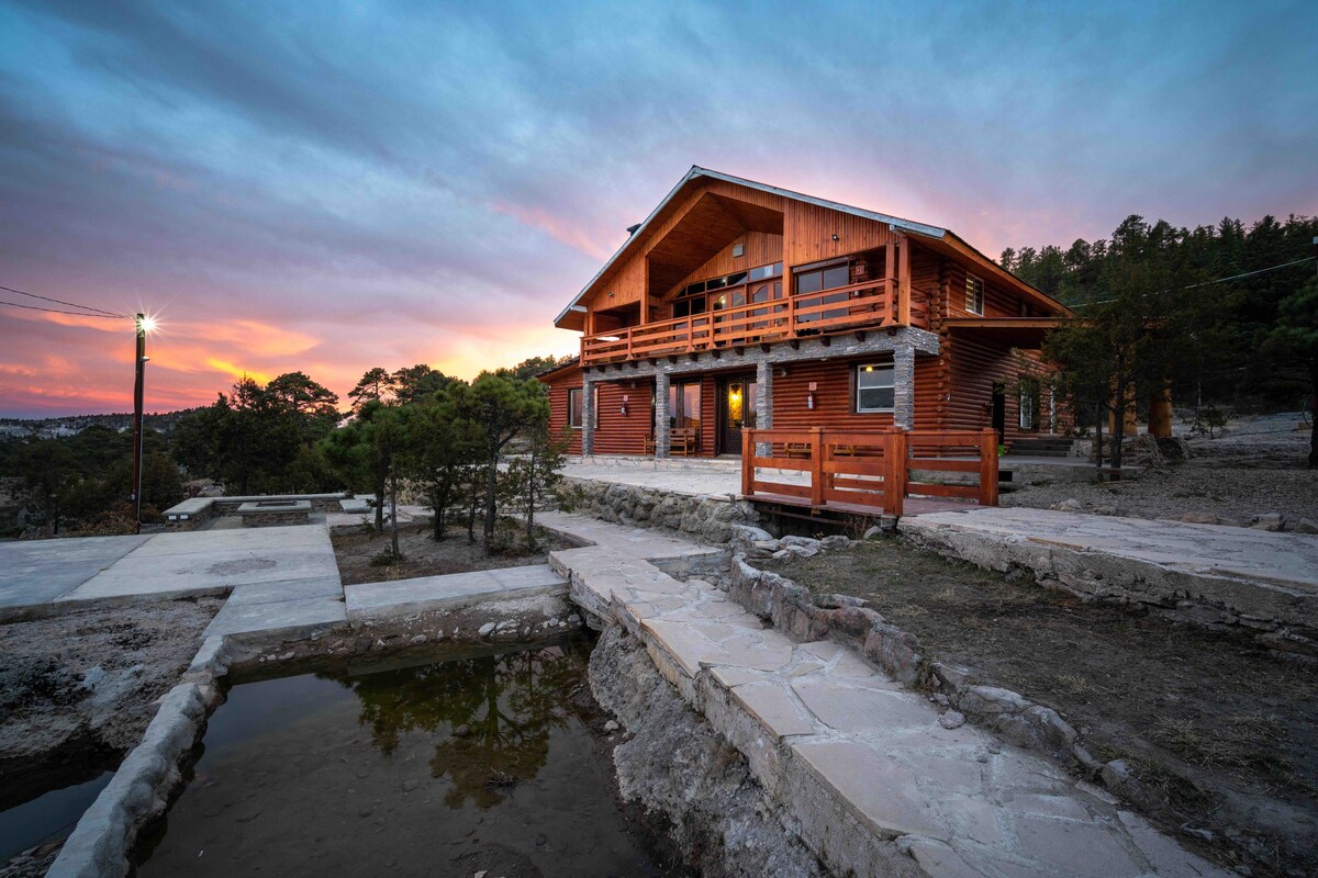 A wooden cabin is set against a vibrant sunset, showcasing a spacious porch with wooden railings. Stone pathways lead to a small pond and surrounding greenery, enhancing the natural landscape. The structure’s large windows reflect the evening light.