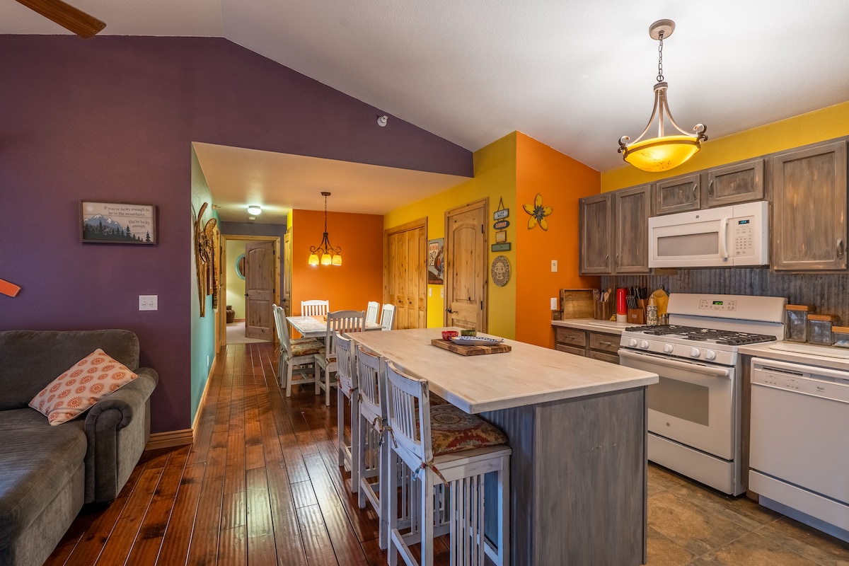 The open kitchen area features colorful walls in shades of orange and purple, highlighting a spacious island with seating for six. A gas stove and microwave are integrated into the cabinetry, accompanied by a dining area illuminated by pendant lights. Warm wood floors create an inviting ambiance.