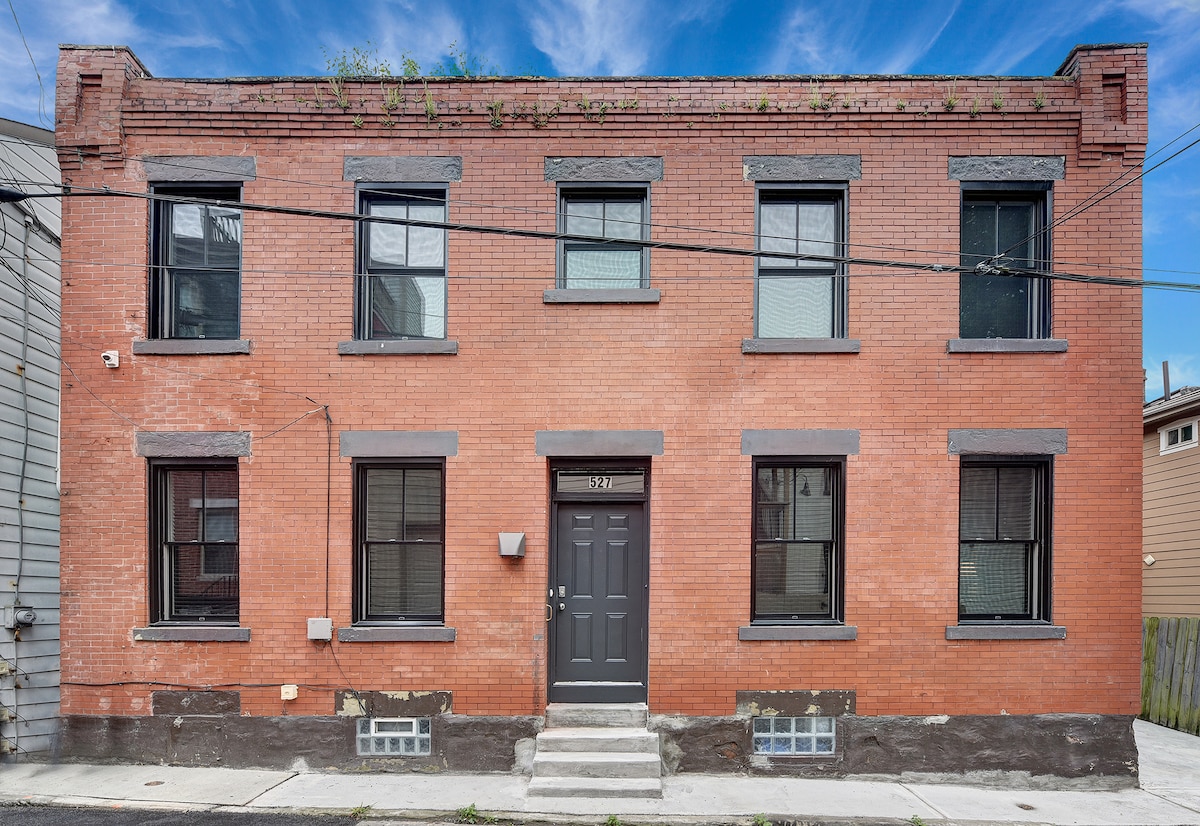 A historic two-story brick building showcases four windows on the upper level, with simple black frames complementing the structure's red exterior. A central door leads to the entryway, framed by subtle architectural details. The exterior is enhanced by greenery sprouting from the top.