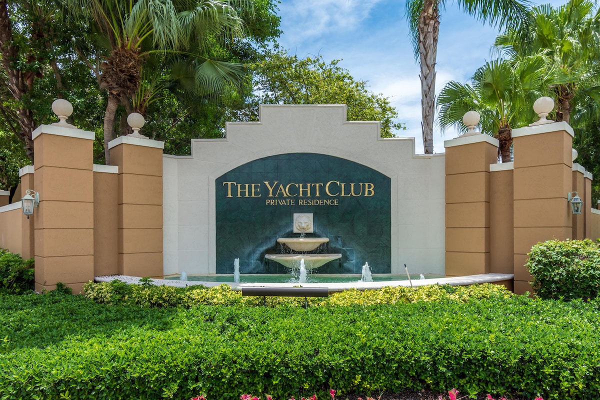 A welcoming entrance to The Yacht Club is displayed, featuring a well-maintained fountain and elegant signage. Lush greenery and palm trees frame the area, contributing to a relaxed ambiance.