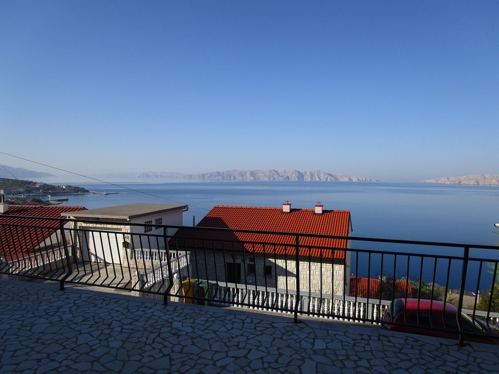 A spacious terrace is showcased, offering stunning views of the sea and surrounding landscape. The red-roofed houses are visible below, along with the glistening water and distant hills. The area is paved with stone, providing ample space for outdoor activities.