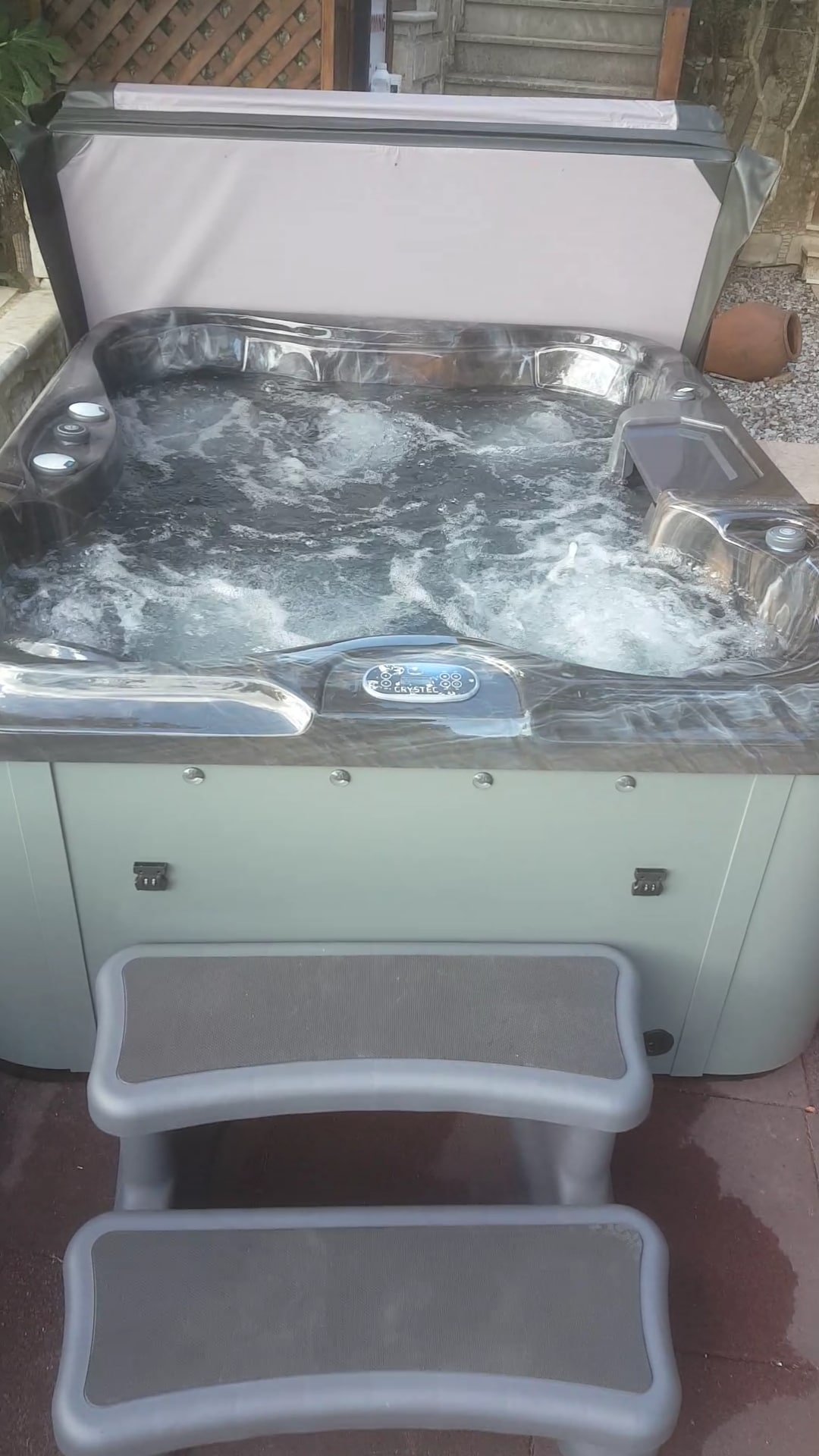 A private hot tub with bubbling water is featured, surrounded by a paved area. Steps lead up to the hot tub, which is designed for five people. The backdrop includes a textured wall and some greenery, enhancing the calm ambiance.