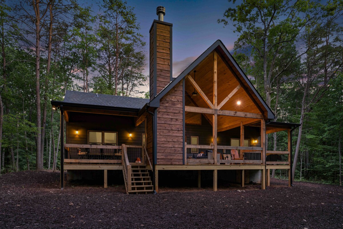 A modern cabin is nestled among trees, showcasing a large front porch with steps leading up to it. The exterior features a mix of wood and stone, with a tall chimney positioned centrally, and warm lights illuminating the porch area.