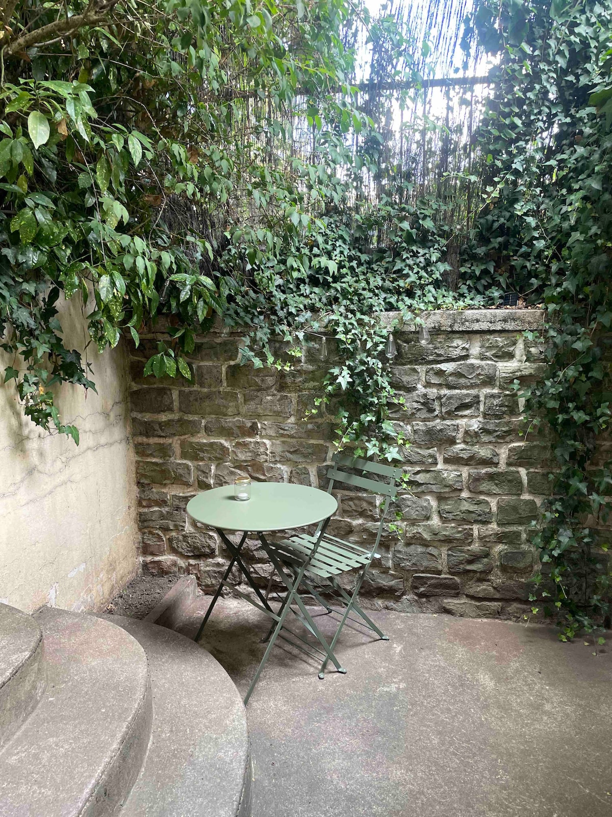 A small outdoor space is featured with a round green table and a folding chair, set against a natural stone wall covered in greenery. The area is shaded by leafy plants, creating a tranquil spot for relaxation.