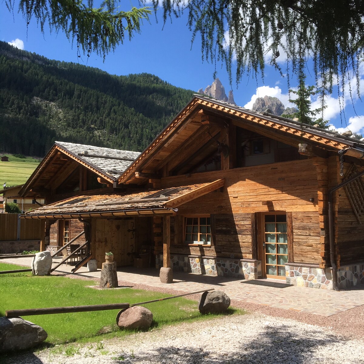 The exterior of the chalet is characterized by rustic wooden construction, complemented by a stone pathway and manicured green lawn. Large windows allow natural light to fill the space, with a backdrop of lush mountains enhancing the tranquil environment.