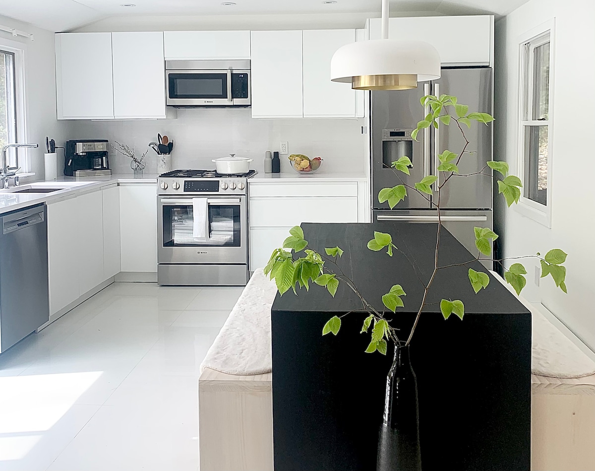 A modern kitchen features sleek white cabinetry and stainless steel appliances, including a gas stove and refrigerator. A black island is enhanced by a minimalist pendant light above, while natural light floods in through large windows, illuminating a simple green plant centerpiece.