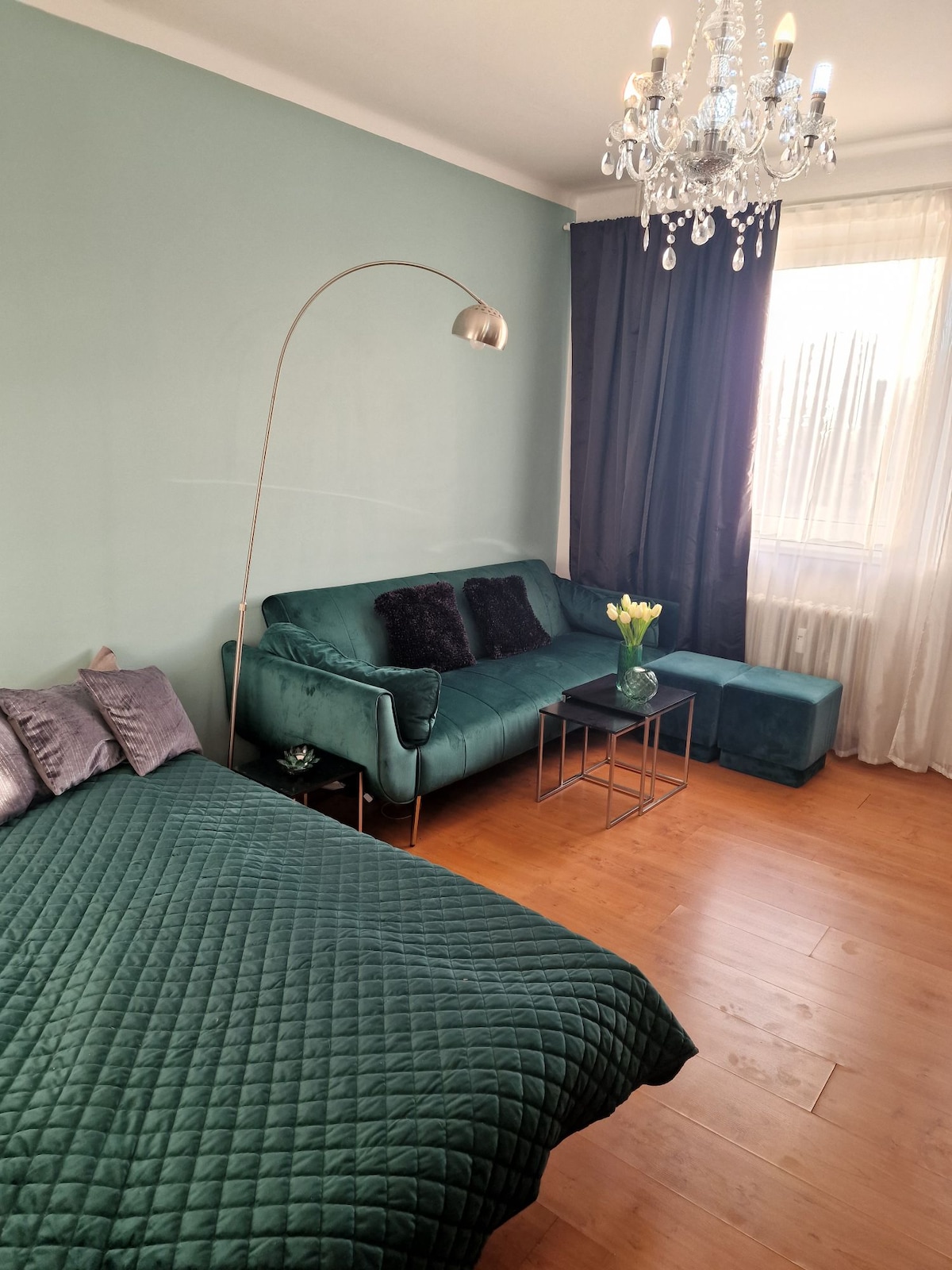 A cozy sleeping area features a plush green bedspread on a spacious bed. A stylish teal sofa is paired with a small side table, adorned with a vase of fresh flowers. Natural light filters through sheer curtains, creating a calming ambience.
