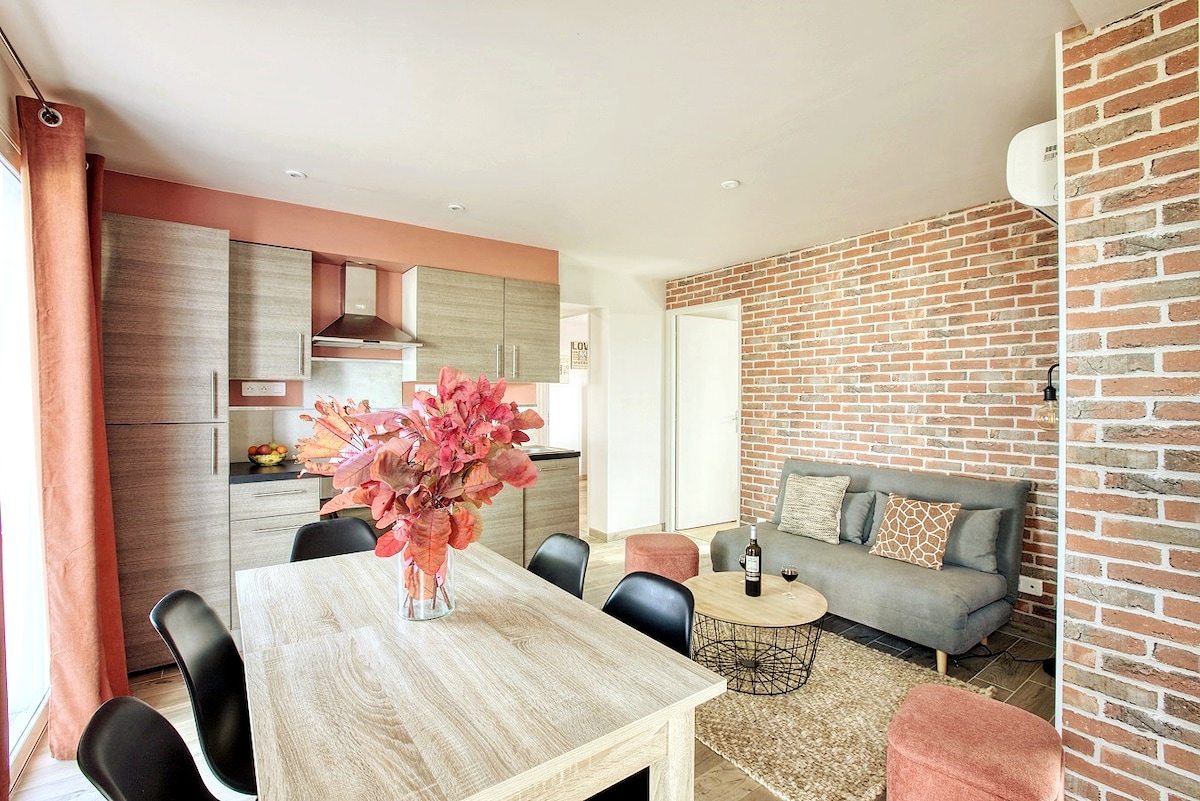 A modern living area features a wooden dining table with black chairs, complemented by a cozy gray sofa. A vibrant floral arrangement is placed at the center of the table, while a textured rug adds warmth to the space. Large windows allow for natural light, enhancing the inviting ambiance.